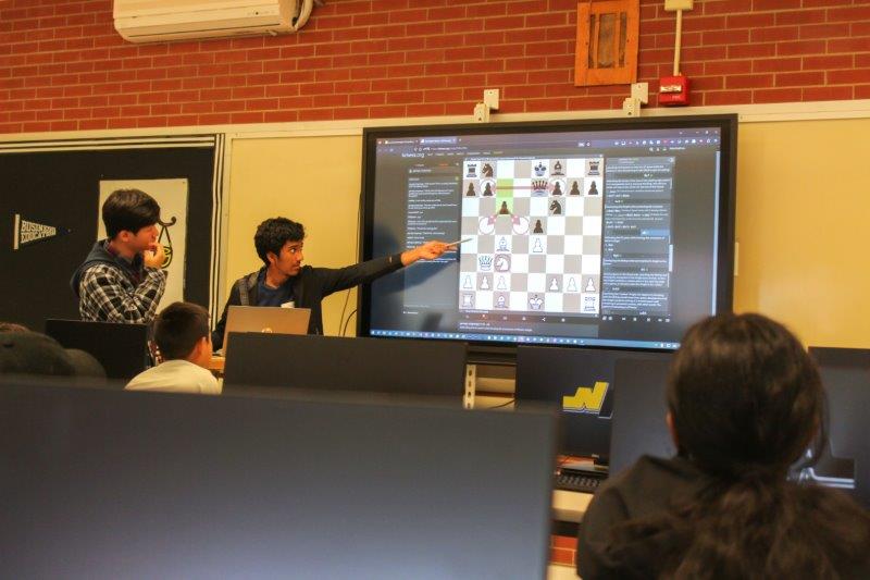 2 teachers are teaching a chess move on a large screen
